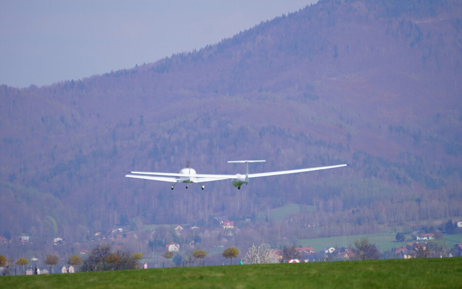 Segelflugzeug SZD-51-1 Junior beim Start über Wiese vor bewaldetem Hügel.
